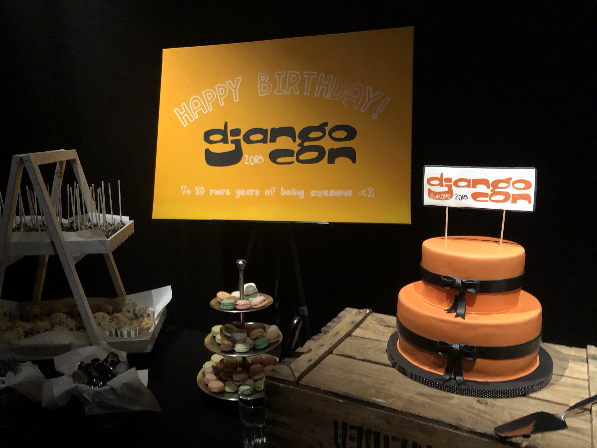 An orange cake on a table, behind it a sign that reads happy birthday djangocon